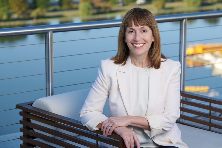 Rebecca Stack, with medium length brown hair and white blazer sits on a chair with water in the bqckground