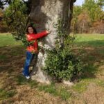 Kristen wearing a red jacket, jeans, and a big smile, hugging a tree and only being able to reach half way around