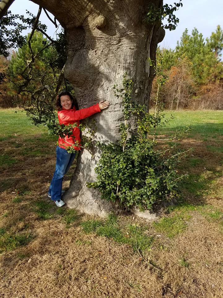 Kristen wearing a red jacket, jeans, and a big smile, hugging a tree and only being able to reach half way around
