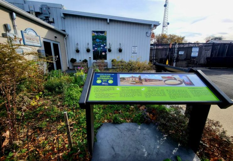A garden grows outside of a one-story building with signage and a mural of a dragonfly and flowers painted on it. The plants are green. An informational display about the park’s commitment to sustainability is in the foreground. 