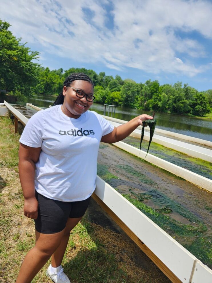  A woman smiles as she holds up a clump of green algae taken from the algal turf scrubber, which consists of water flowing over long white panels next to her. A body of water and green trees are in the distance.