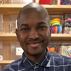 A man in a blue button-down shirt smiles in front of a bookcase with clear containers of colorful supplies.